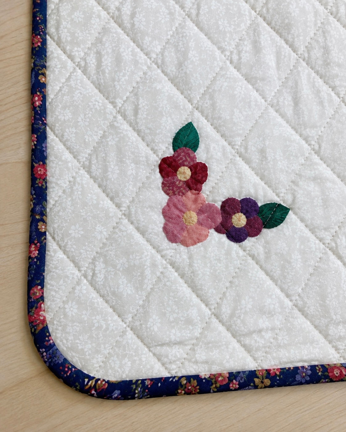 A close-up of a quilted baby quilt with a floral embroidered motif in pink, red, purple, and green near the center. The fabric has a dark navy floral border and is laid on a light wood surface.