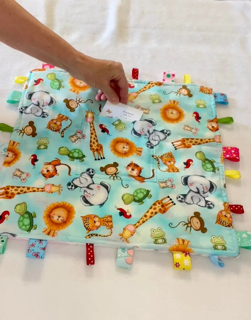 A baby sensory lovey blanket with a light blue background and colorful safari animal print, featuring a hand holding a small paper tag on the fabric, also good for gift cards, special baby note.