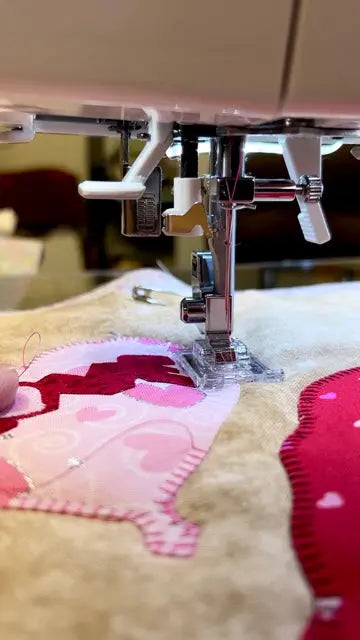 Close-up of a sewing machine needle and presser foot stitching a pink and red patchwork quilt on beige fabric.
