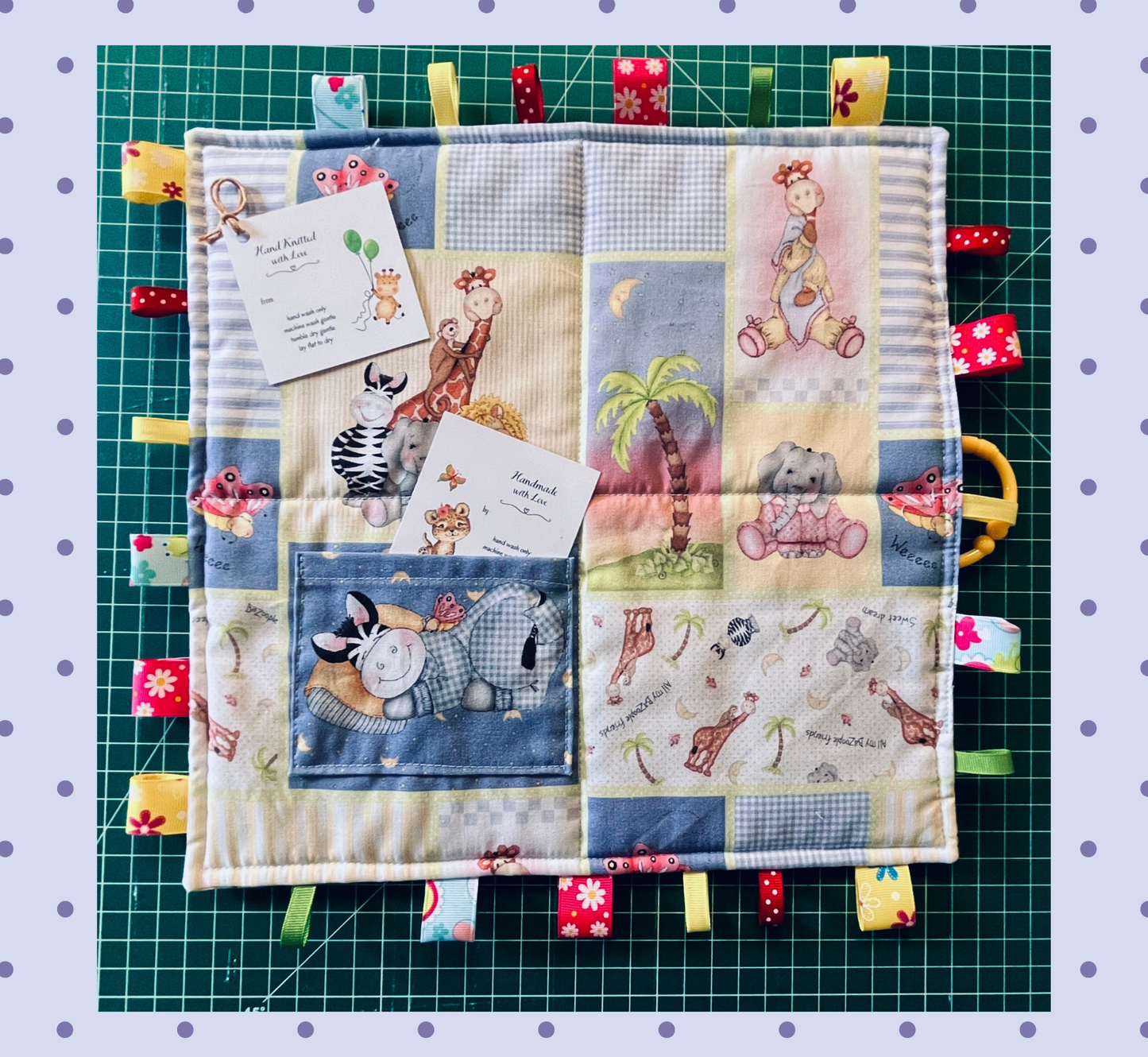 A square quilted baby sensory blanket with animal patches and colorful fabric tabs, displayed on a green cutting mat with scissors and tags. Includes pocket for gift card