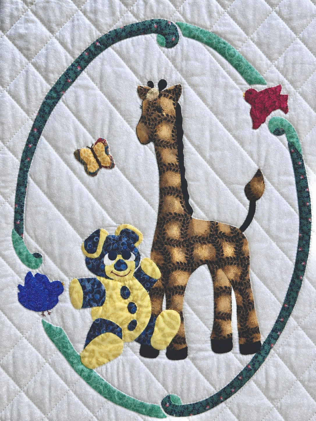 A close-up of a quilted fabric featuring a giraffe and teddy bear applique design inside a green oval border, with butterflies and flowers on a white quilted background.