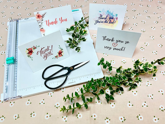 Set of greeting cards with 'Thank you' messages on a floral-patterned surface with scissors and greenery.